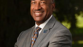 Chancellor Gary S. May portrait stands with his arms crossed, smiling, in a grey suit in front of a dark green background