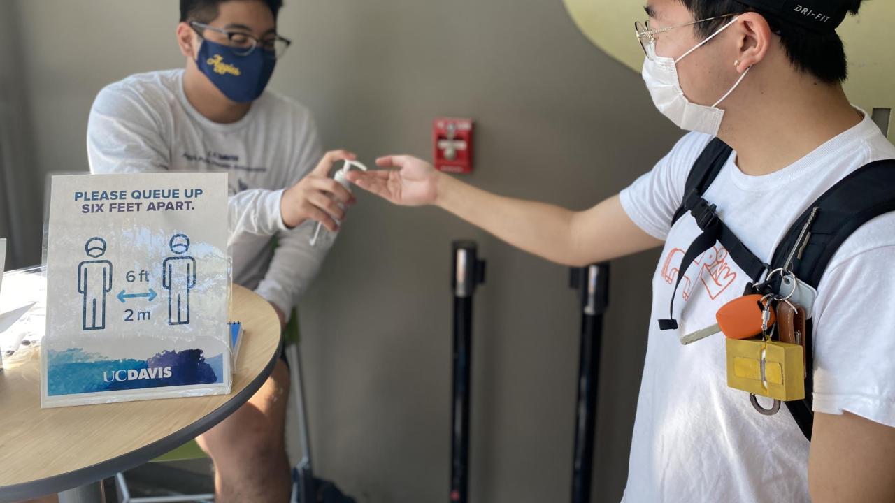 Masked public health ambassador providing hand sanitizer to student.