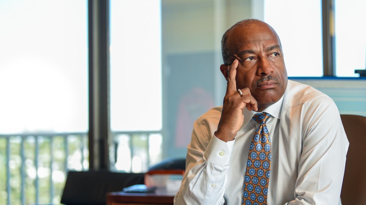 Chancellor May sitting at desk with his right hand on his temple, looking to the left outside a window