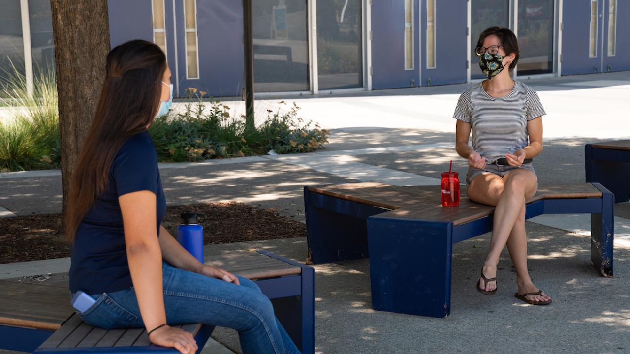 Two UC Davis students sitting socially distanced while wearing face coverings on benches.