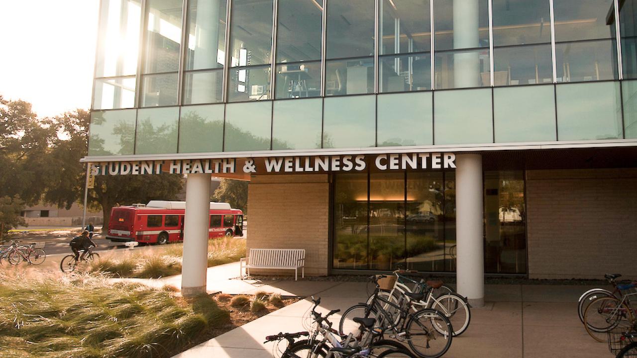 Student Health and Counseling Services Center building with bike racks, bus and person riding a bike