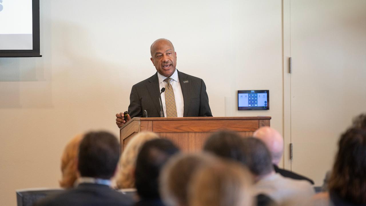 A speaker stands at a podium addressing an audience in a conference setting.