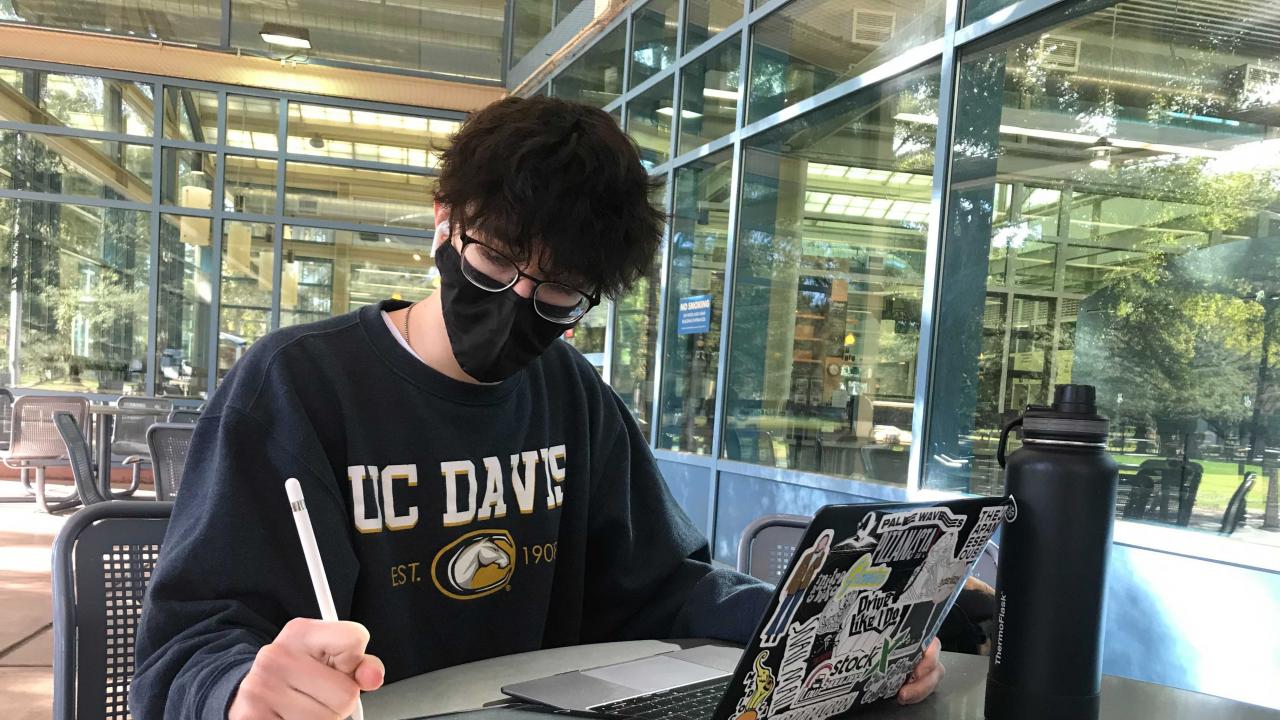 Student with mask working on laptop and tablet with UC Davis sweatshirt