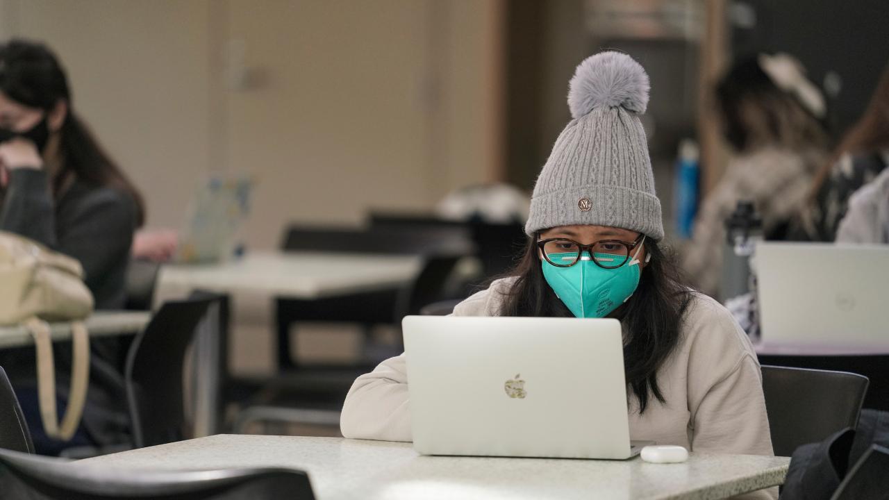 Student behind laptop with mask and hat.