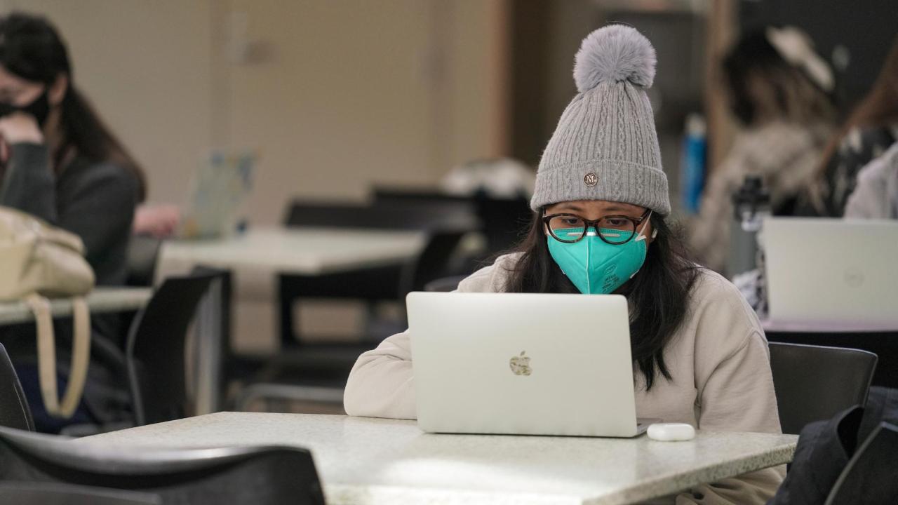 Student behind laptop with mask and hat.