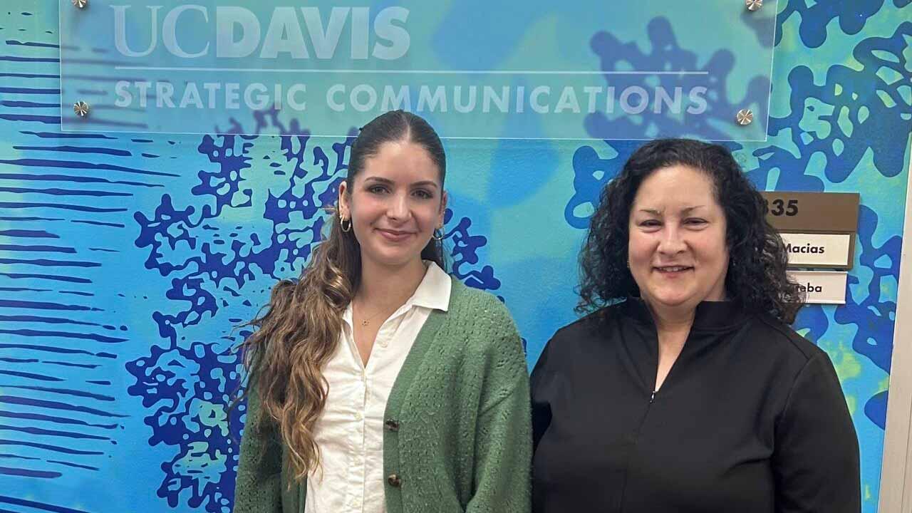 Two people pose for photo in front of blue wall with plaque: UC Davis Office of Strategic Communications