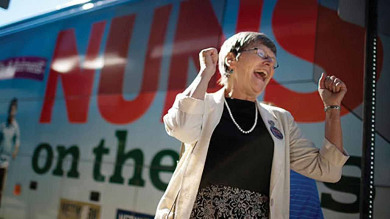 Sister Simone raises arms in jubilation outside bus with "Nuns on a Bus" banner