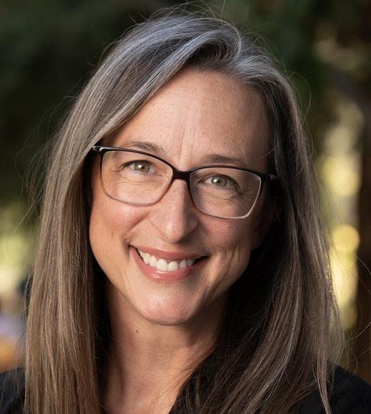 Smiling woman with glasses and long hair stands in front of trees in softly lit outdoor setting