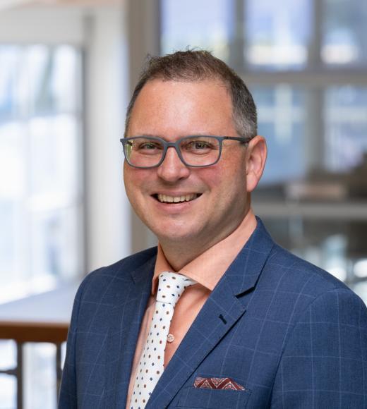 Headshot of smiling man in blue suit and glasses, peach shirt and polka-dot tie, office background