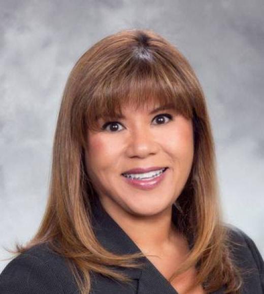 Professional headshot photograph of smiling woman with shoulder-length brown hair, gray background
