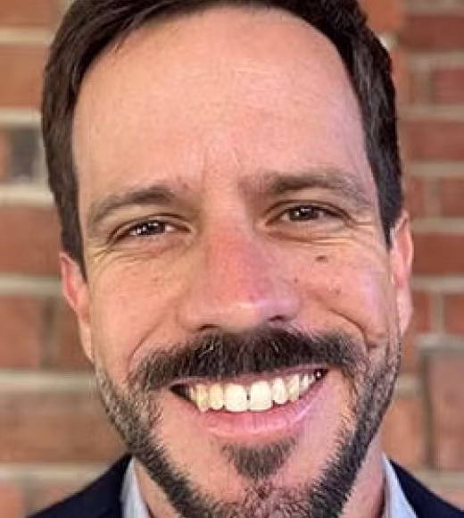 Smiling male headshot with dark hair and goatee, wearing a blazer against a brick wall background