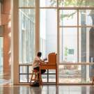 Two people sitting at separate desks spaced far apart in the library adjacent to the courtyard windows.