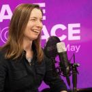 Woman (Professor Amanda Guyer) with long brown hair smiling into studio microphone, purple 'Face to Face' backdrop