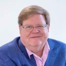 Smiling older man wearing glasses, blue blazer and pink shirt against white background