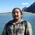 Young man with backpack smiles by a body of water and rocky mountains.