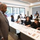 A group of students dressed in black hoodies and a white sweater show Chancellor May dressed in a brown blazer jacket informational graphics on a white table. 
